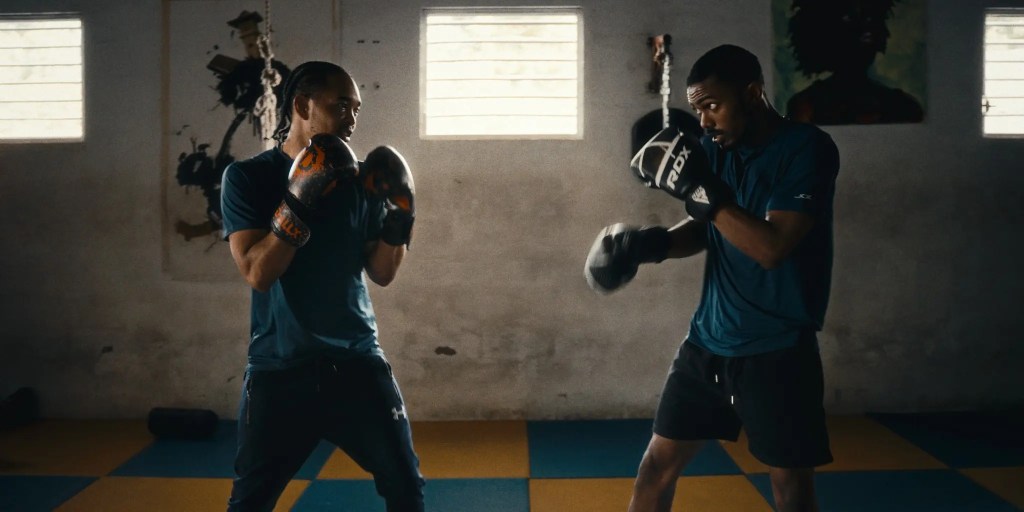 Two male boxers in training stance, wearing gloves, engaged in a sparring session in a gym setting with colorful mats and windows visible.