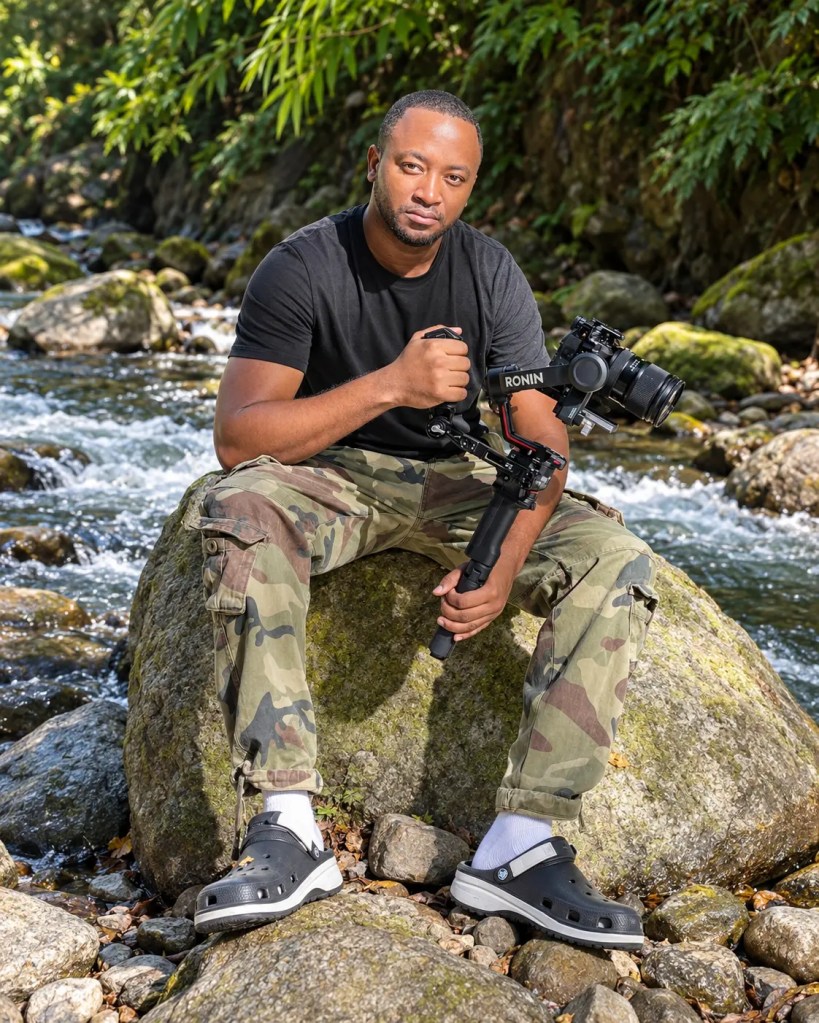 A man sitting on a rock by a stream, wearing camouflage pants and a black shirt, holding a camera stabilizer with a camera attached.