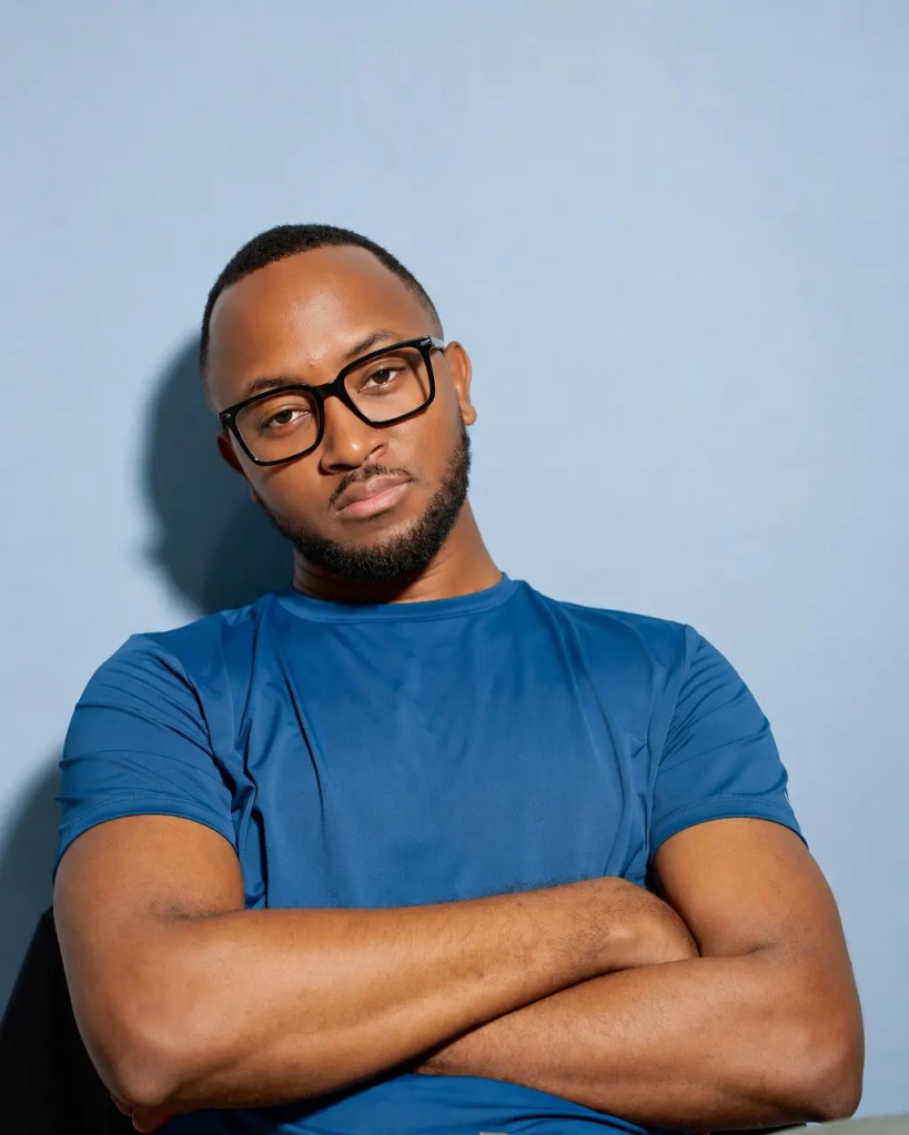 A man wearing glasses and a blue athletic shirt, sitting with his arms crossed, against a light blue background.