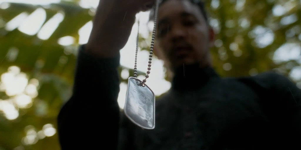 A person holding a dog tag necklace in a natural environment, with trees blurred in the background.
