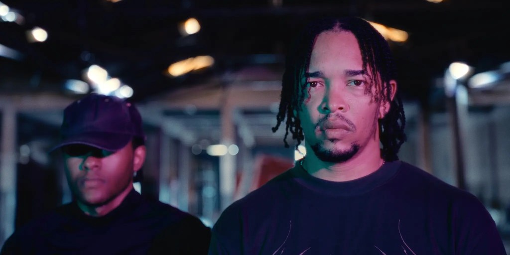 Two men standing in a dimly lit industrial space, with one man in front displaying a serious expression and the other behind him wearing a cap.