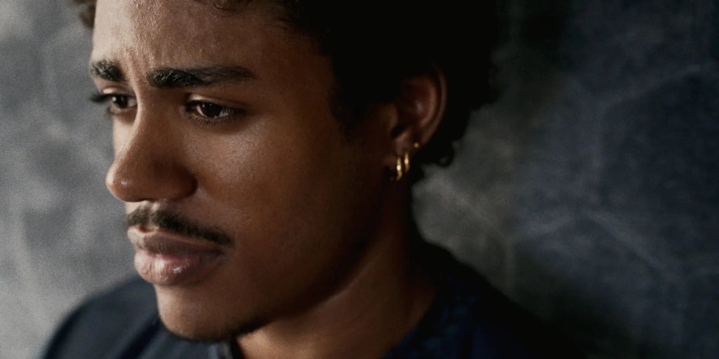 Close-up of a young man with curly hair, featuring a thoughtful expression and wearing small gold hoop earrings.